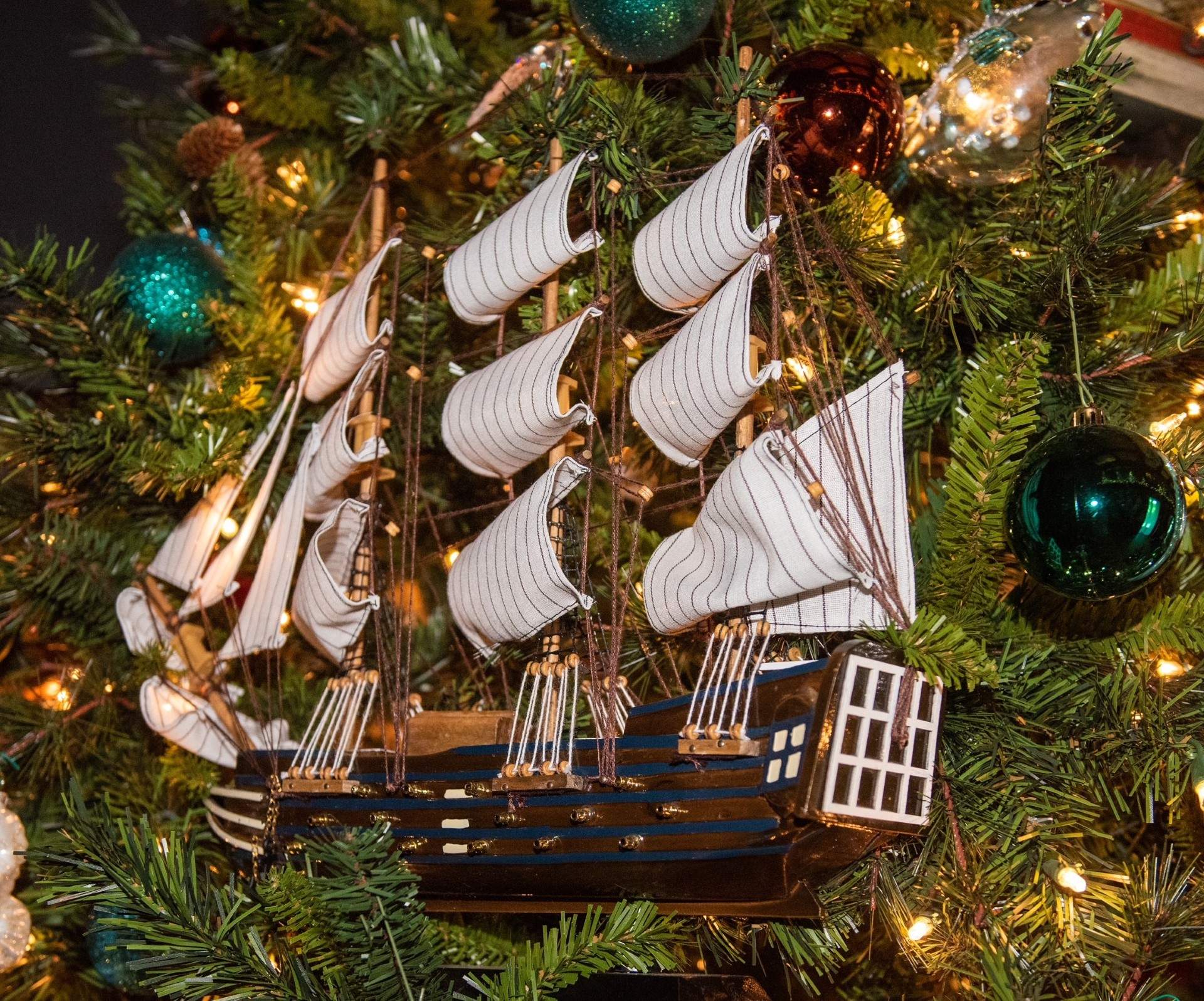A detailed model of a wooden sailing ship ornament with multiple white striped sails hangs on a decorated Christmas tree. The tree is adorned with green, teal, and bronze ornaments, pine needles, and warm white lights surrounding the ship.
