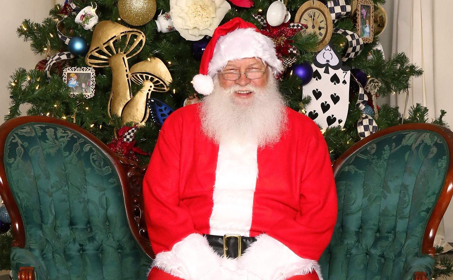 Santa Clause wearing his red and white Santa suit and hat sits on a green sofa in front of a Christmas tree and smiles.