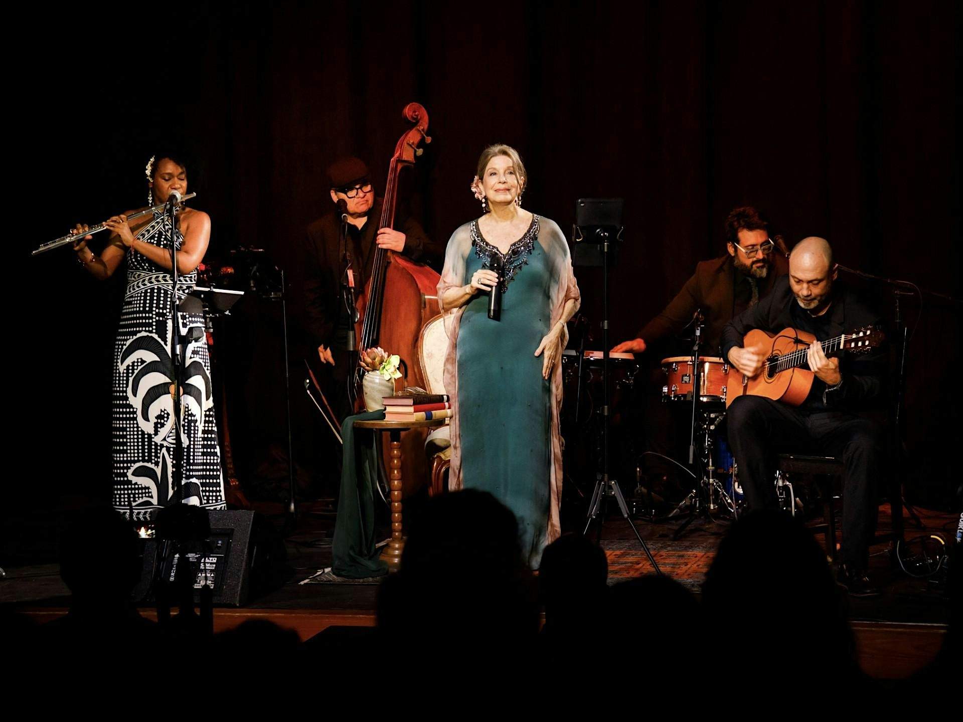 A woman in a black and white dress playing the flute, a man playing the standing bass, a man playing the drums, a man playing an acoustic guitar, and a woman in a green dress singing into a microphone on stage.