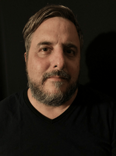 A man in a black shirt against a black background with short brown hair and a short brown beard looking into the camera.