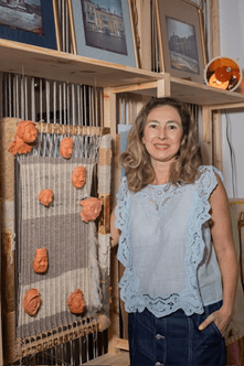 A woman with dirty blonde hair wearing a blue tanktop and jean skirt stands smiling at the camera next to an art piece.