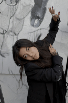 A woman with long black hair looks seriously into the camera as she performs an interpretive dance.