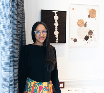 A woman with long black hair and glasses dressed in a black long-sleeve shirt and a colorful skirt stands in front of abstract artworks.