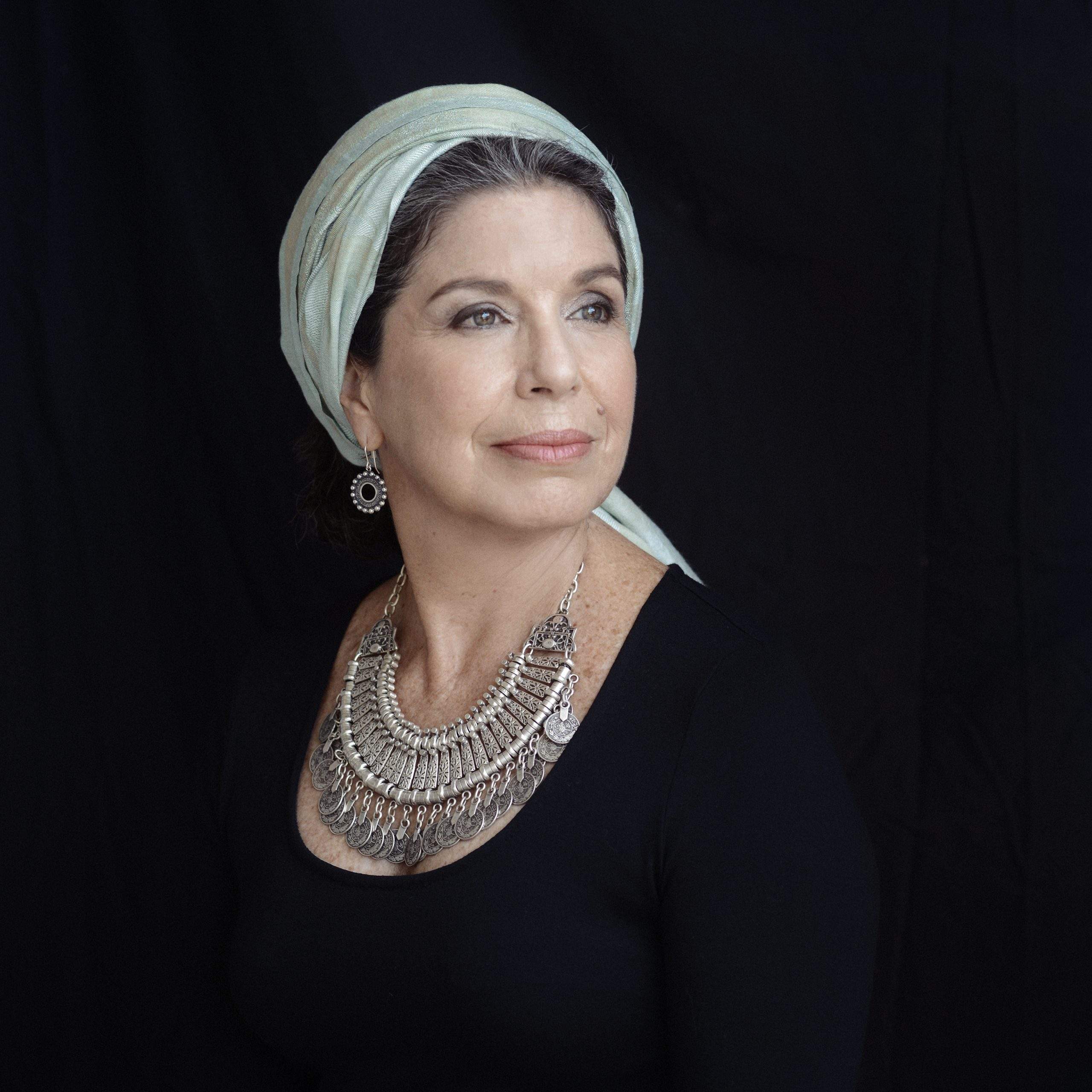 A person is posed against a dark backdrop, wearing a light-colored headscarf wrapped around the head, a dark long‑sleeved top, a large layered metallic necklace with ornate detailing, and a dangling circular earring. The lighting highlights the accessories and upper torso.