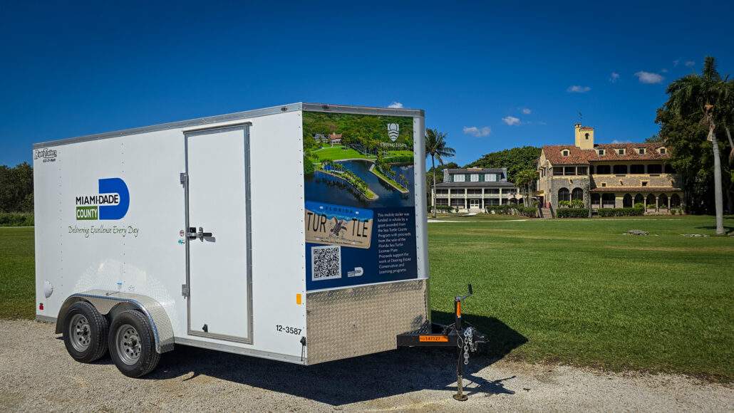A trailer with the Miami-Dade County logo on the side and a graphic featuring the Boat Basin, Deering Estate Logo, and TURTLE license plate on the front parked in front of the historic homes and sprawling main lawn of Deering Estate.