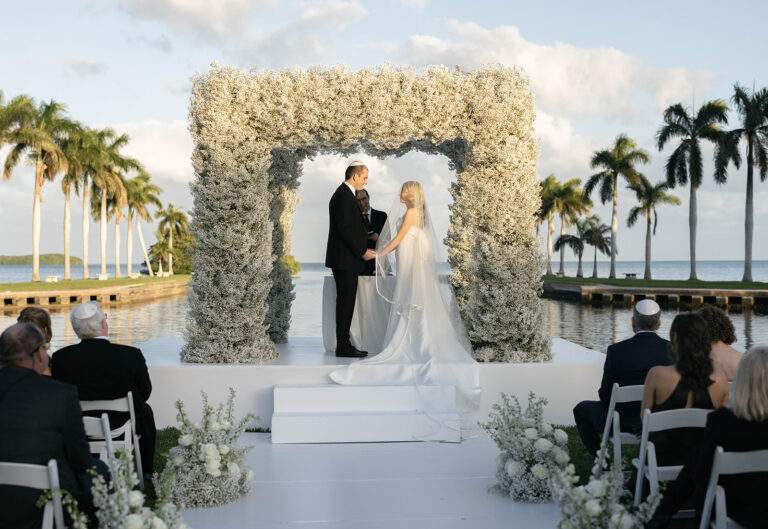 A groom and bride hold hands and look lovingly at one another at the altar of their wedding ceremony in front of a large floral arch.