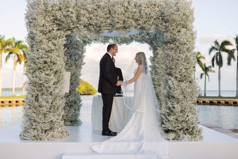 A groom and bride hold hands and look lovingly at one another at the altar of their wedding ceremony in front of a large floral arch.