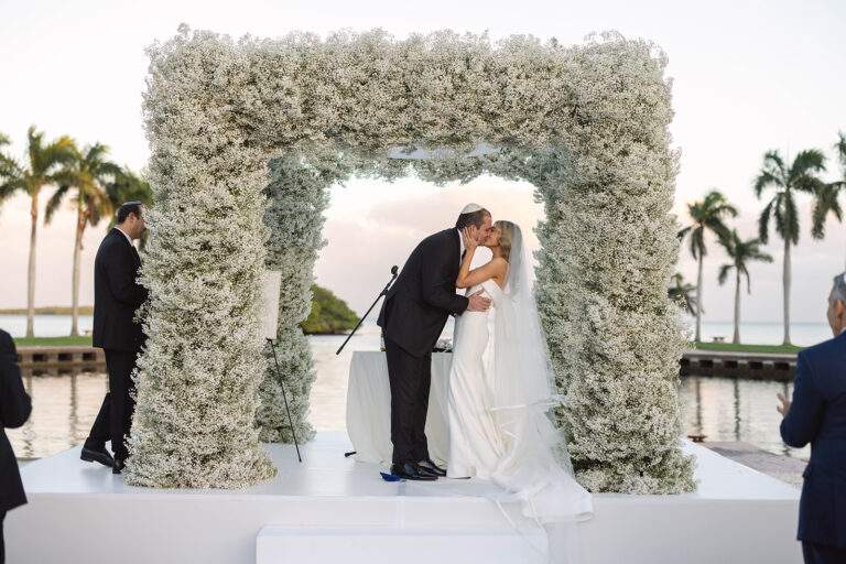 A bride and groom kissing at the altar of their wedding ceremony in front of a large floral arch.