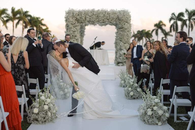 A groom dips his bride as he kisses her in the aisle during their wedding ceremony, surrounded by floral decorations and their loved ones.
