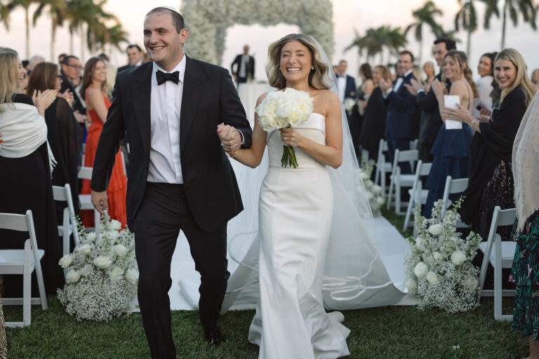 A bride and groom joyfully strut down the aisle together during their wedding ceremony, surrounded by floral decorations and their loved ones.