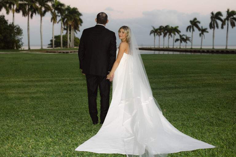 A photo of a groom and bride on the Main Lawn at Deering Estate with a view of Biscayne Bay. The groom, wearing a black tux, is faced away from the camera looking out to the water, and the bride is looking back at the camera as she holds the groom's hand.