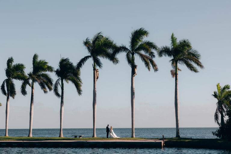 View of a bride and groom near the water on the jetty with palm trees at Deering Estate.