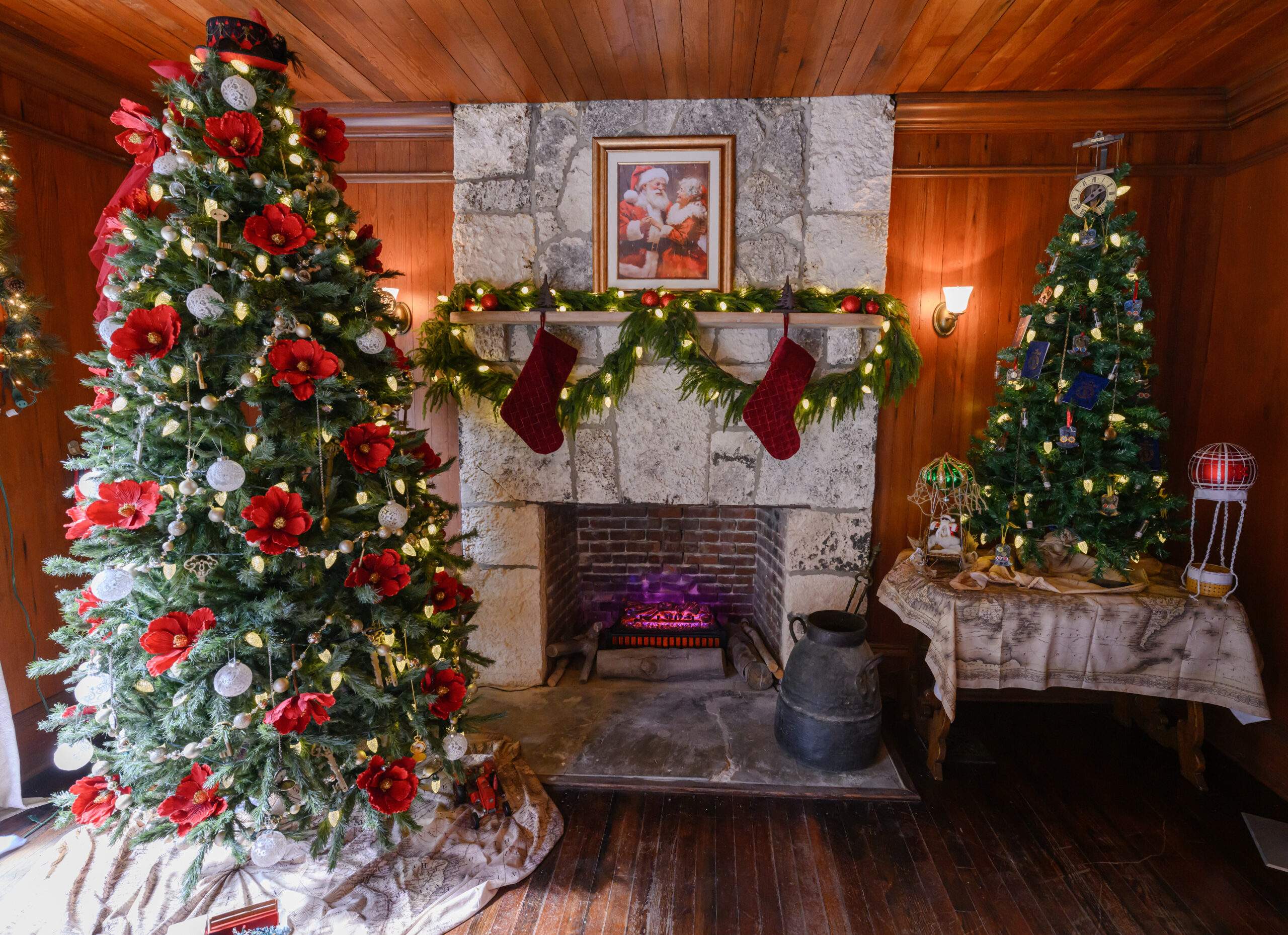 The fireplace area of the dining room in the Richmond Cottage decorated for the holidays. A photo of Mr. and Mrs. Clause hangs above the fireplace. To the left of the fireplace is a Christmas tree with poinsiettas and baubles. To the right of the fireplace is a smaller Christmas tree on a table with large white lights.