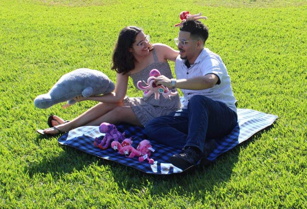 A couple sitting on a Deering Estate-branded picnic blanket on the Main Lawn playing with a manatee and octopus plushies.