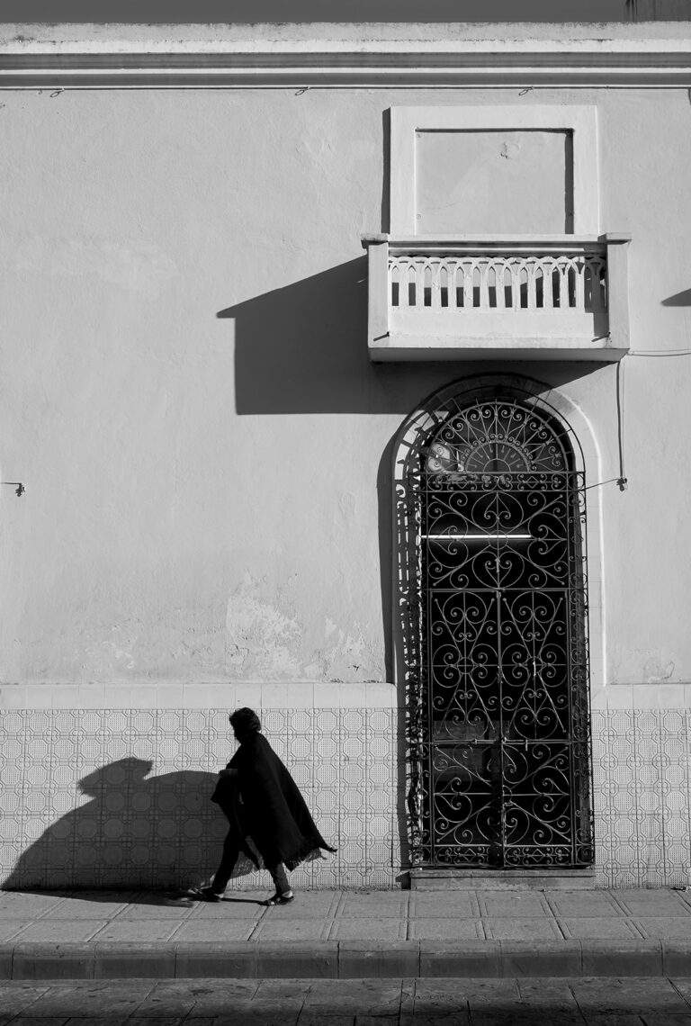 A photograph on a person in a black shawl walking on a sidewalk next to a tiled buidling.