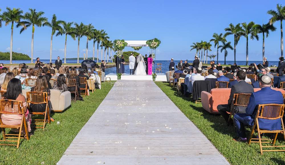 A bride and groom stand at the altar in front of the Boat Basin. Lining the aisle are seats filled with family and loved ones.