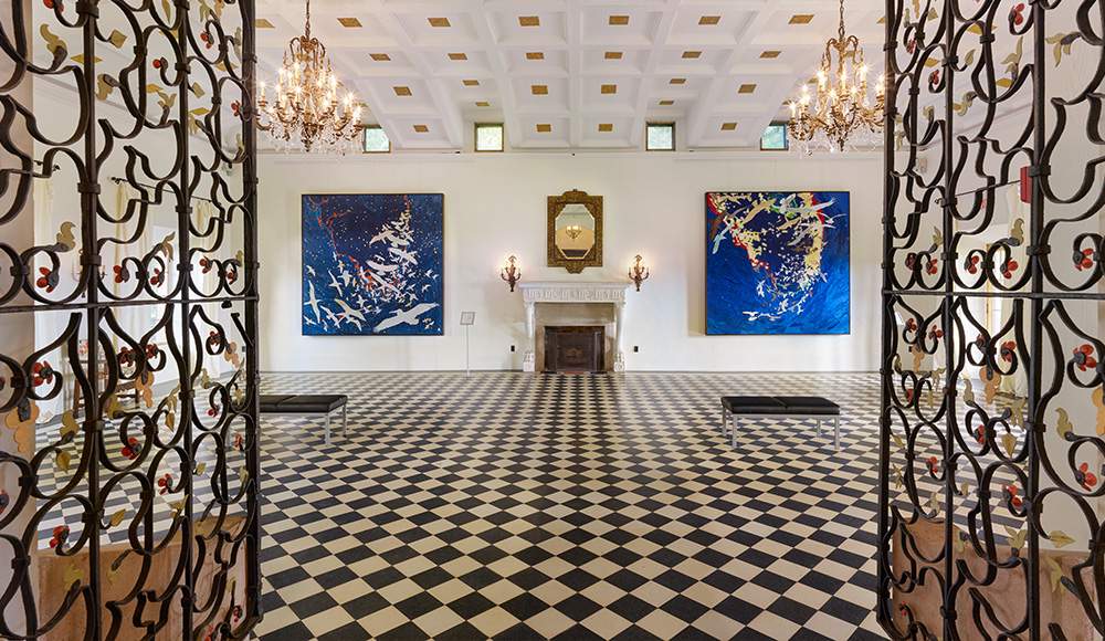 The Great Hall inside the Stone House. It has a white and black checkered floor and white walls with a fireplace and mantel in the center on the far wall. The entrance has wrought-iron gates with metal floral decoration.