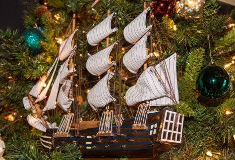 A detailed model of a wooden sailing ship ornament with multiple white striped sails hangs on a decorated Christmas tree. The tree is adorned with green, teal, and bronze ornaments, pine needles, and warm white lights surrounding the ship.