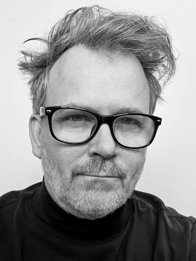 A black and white image of a man with short grey hair and black glasses wearing a black turtleneck shirt looking at the camera. This is artist Barron Sherer.
