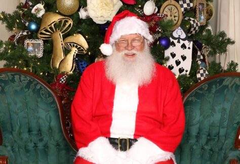 Santa Clause wearing his red and white Santa suit and hat sits on a green sofa in front of a Christmas tree and smiles.