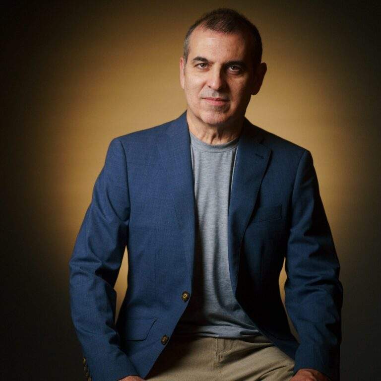 A portrait of artist Jorge Gonzalez Graupera. He is wearing a blue suit jacket over a greyish-blue shirt and khaki pants and is sitting on a stool.