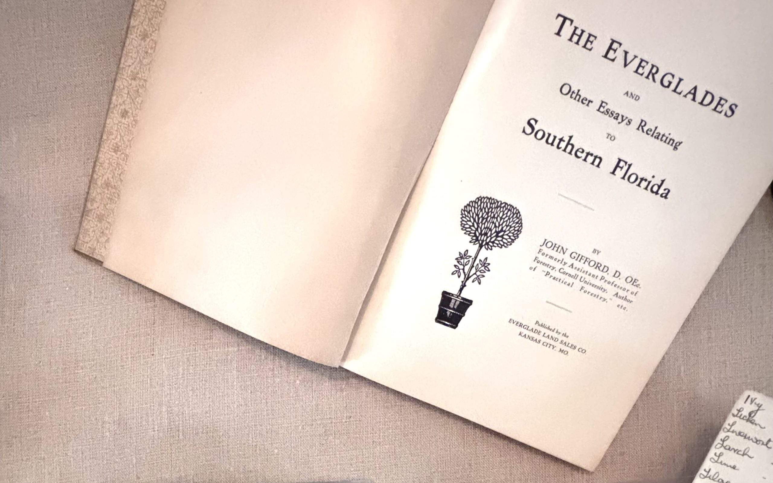 An open book rests on a light, textured fabric surface. The left page is blank, while the right page shows a title page with printed text reading “The Everglades and Other Essays Relating to Southern Florida.” Below the title is a small black illustration of a potted, round-canopied tree. Additional text on the page provides the author’s name and publishing information in smaller print. In the lower-right corner of the image, a separate small piece of paper with handwritten notes is partially visible.