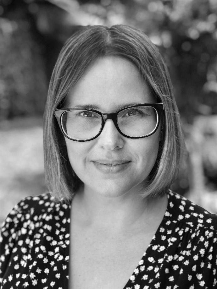 A black and white image of a woman with short hair and glasses wearing a floral top looking at the camera. This is artist Lissette Schaeffler.