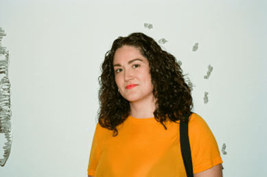 A woman with dark brown curly hair wearing bright coral lipstick and a bright mustard colored shirt smiles at the camera.