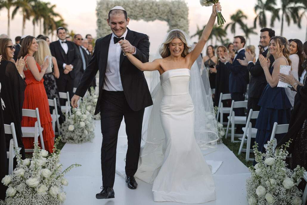 A bride and groom joyfully strut down the aisle together during their wedding ceremony, surrounded by floral decorations and their loved ones.