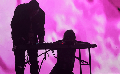 Two people stand at a table with an audio mixer set up in front of pink/purple projected abstract visuals.