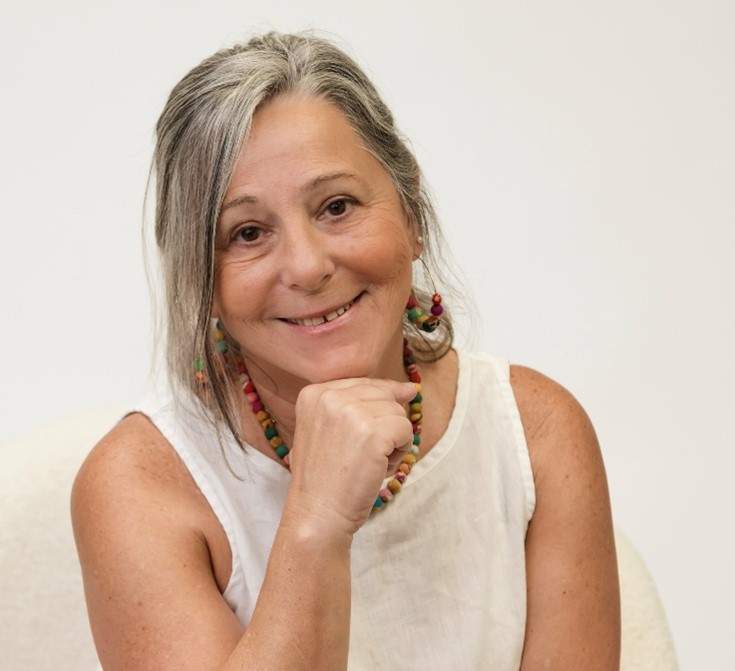 A woman with grey long hair wearing a white tank blouse and colorful earrings and necklace resting her head on her fist and smiling at the camera. This is artist Debora Rosental."