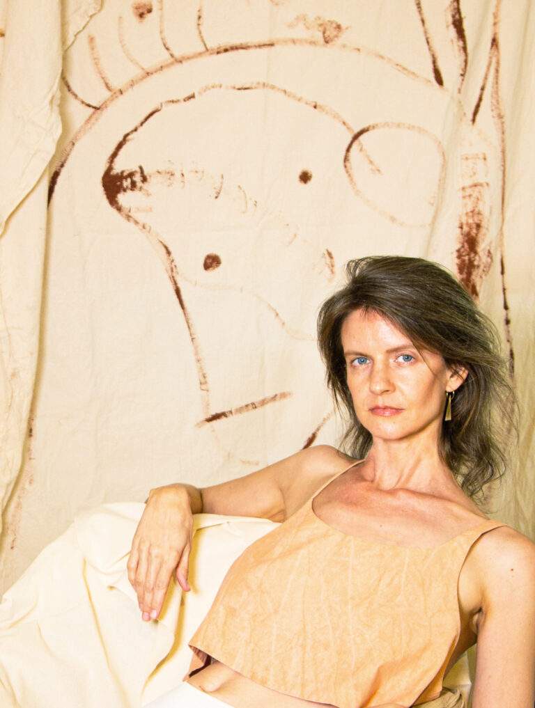A woman with medium-long brown hair in an orange tank top looks at the camera and lounges on a seat with her arm at the top of the chair. This is artist Emily Peters.