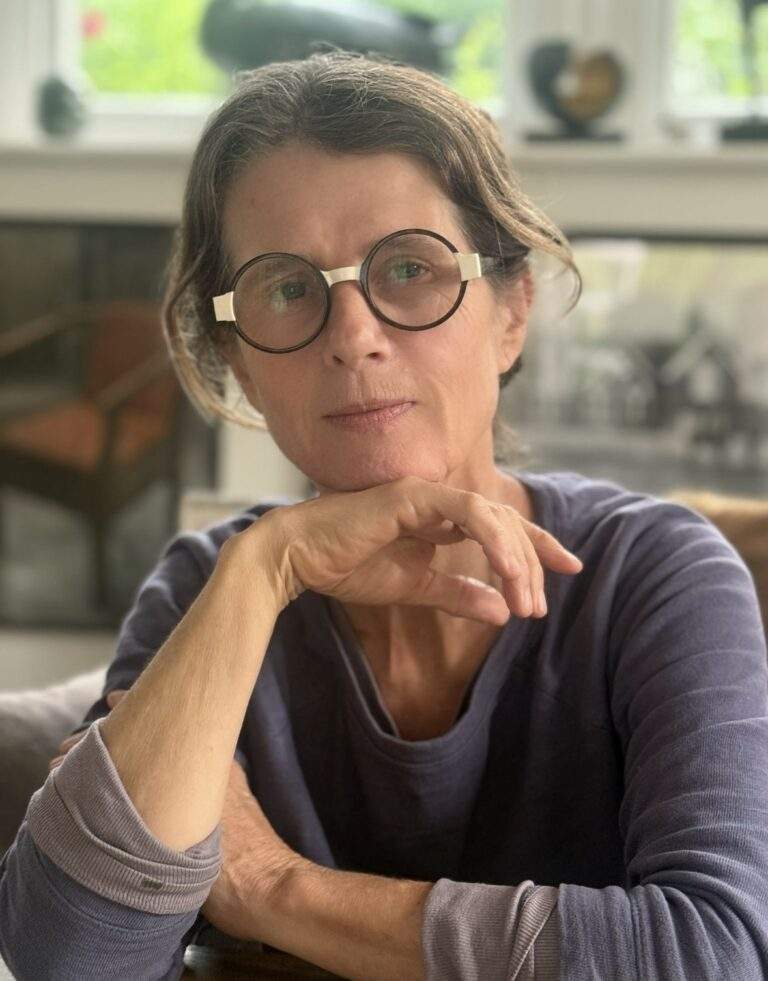 A woman with brown hair in a ponytail with black circular glasses with her head resting on the back of her hand. This is artist Deryn Cowdy.