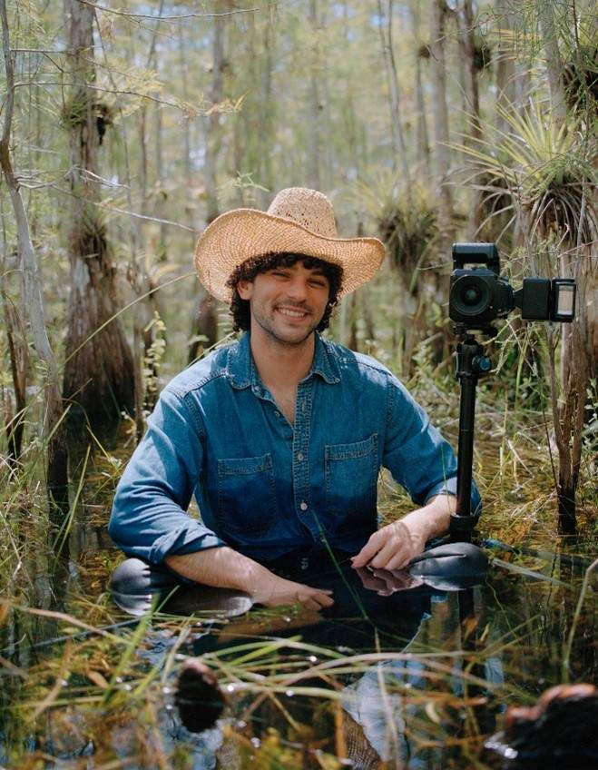 A man in a straw hat and denim button down shirt sits with a camera in the water and is surrounded by trees in the Florida Everglades.