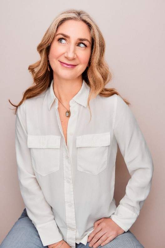 A blonde woman wearing a white button down shirt and jeans sitting on a stool and looking off to her right. This is writer Rebekah Shoaf.