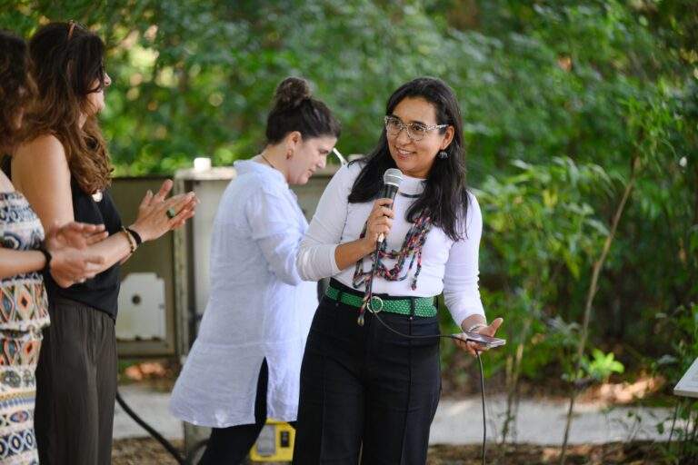 Curator Dainy Tapia. A woman with black hair, mulicolored glasses, white long-sleeve shirt, black pants, green belt, smiling and holding a microphone.