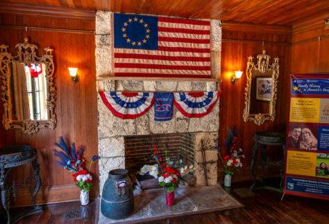 A cozy, wood paneled room features a stone fireplace decorated with patriotic American bunting in red, white, and blue. Above the mantel hangs an early American flag with a circle of thirteen stars. Floral arrangements in matching colors sit on either side of the hearth, along with rustic metal containers and tools. Two ornate mirrors flank the fireplace, and warm wall sconces provide soft lighting. A tall informational display about family history stands to the right.