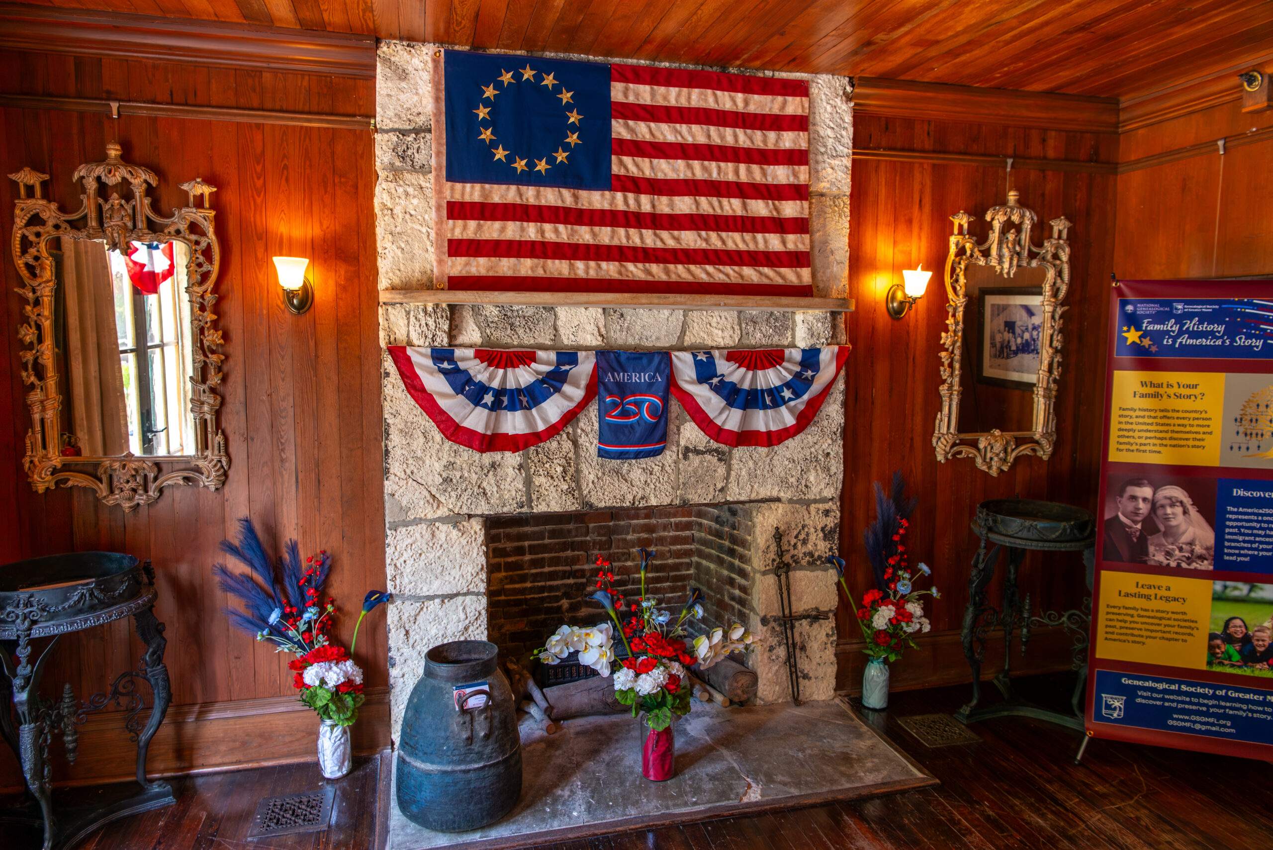 A cozy, wood paneled room features a stone fireplace decorated with patriotic American bunting in red, white, and blue. Above the mantel hangs an early American flag with a circle of thirteen stars. Floral arrangements in matching colors sit on either side of the hearth, along with rustic metal containers and tools. Two ornate mirrors flank the fireplace, and warm wall sconces provide soft lighting. A tall informational display about family history stands to the right.